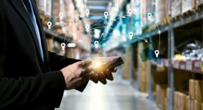 A person holds a smartphone in a warehouse, with a holographic global map showing interconnected shipping routes and location pins, symbolizing modern logistics and supply chain management.
